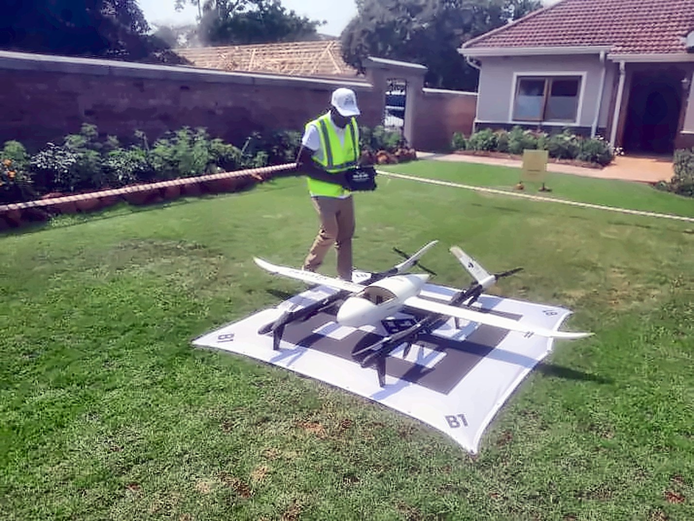 An operator in a high-visibility vest standing beside a white fixed-wing delivery drone on a landing mat on a lawn