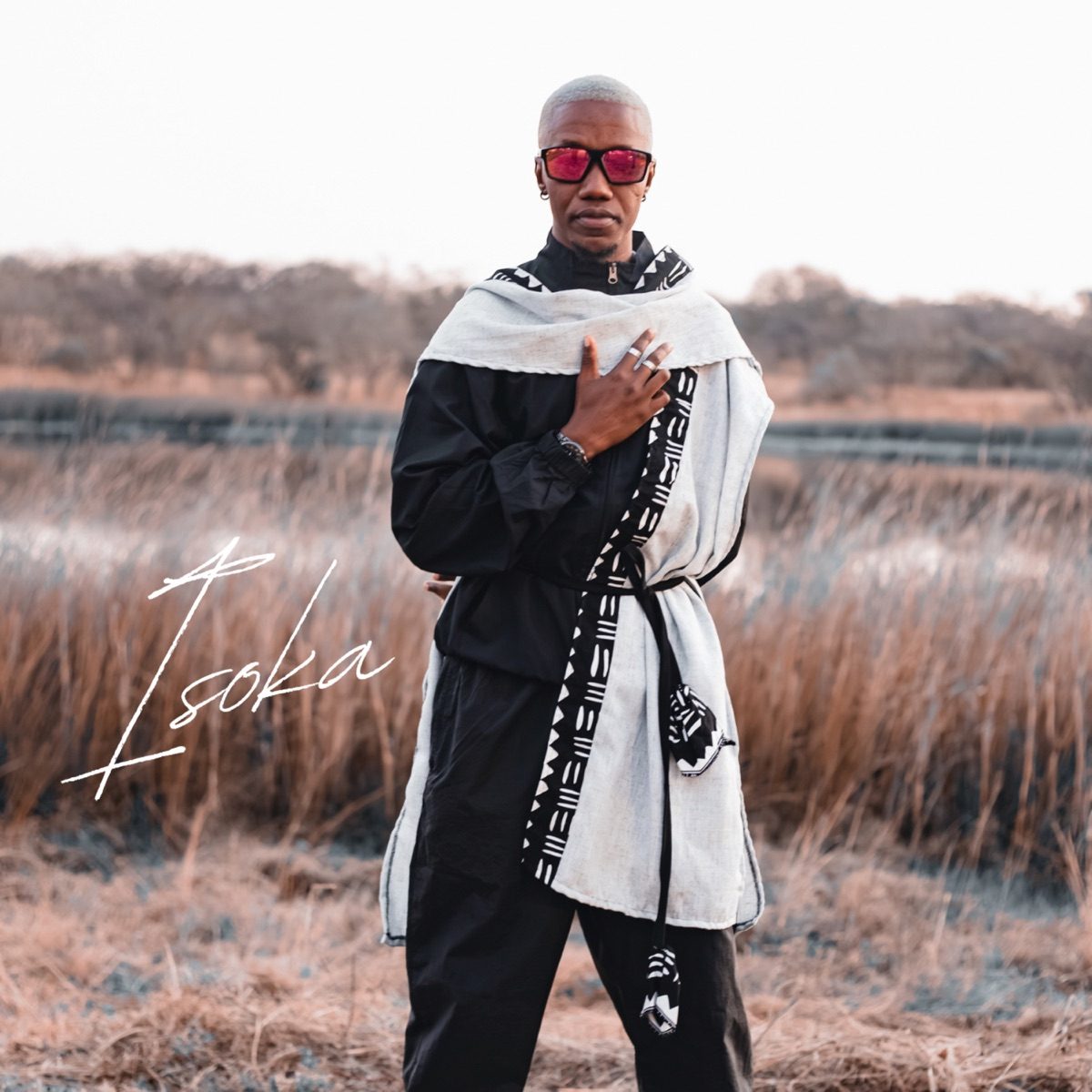 Cover art for Isoka showing a man in red sunglasses and a black and white patterned wrap standing in a dry grass field, with the word Isoka across the image
