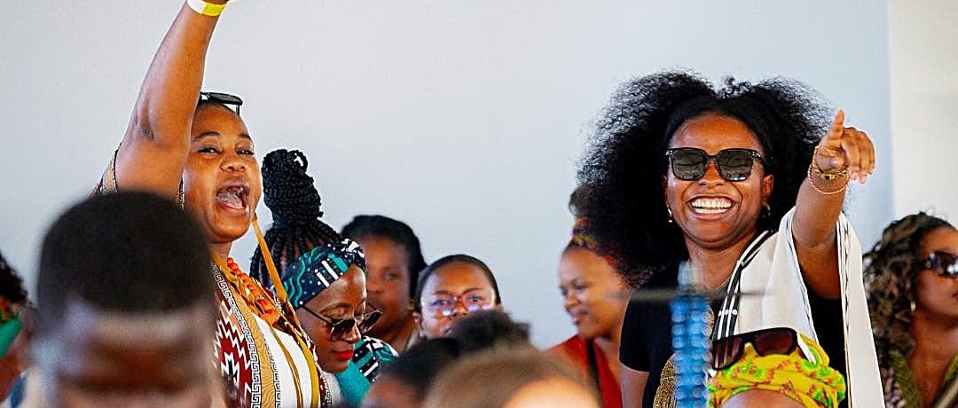 A crowd of women in African print and sunglasses cheering and pointing at a daytime Soke party