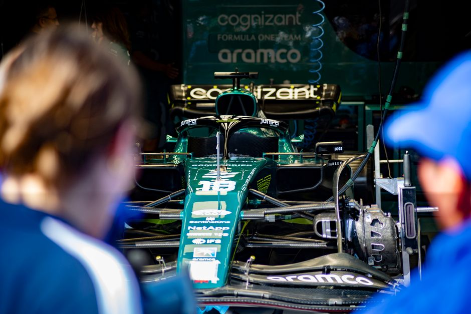 An Aston Martin Aramco Formula 1 car in its garage, seen between two blurred figures in the foreground