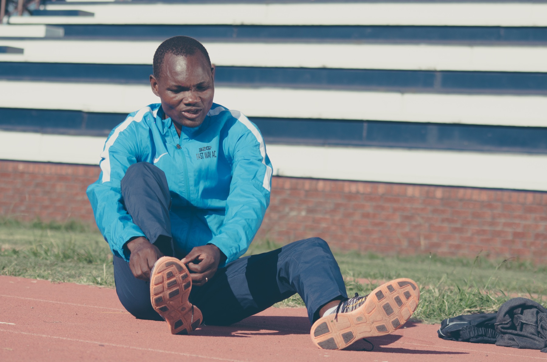 Stephen Muzhingi in a blue tracksuit jacket sitting on a running track, adjusting his running shoe
