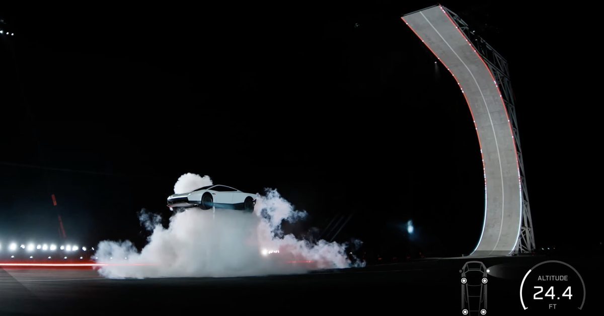 A white Tesla Roadster airborne above a cloud of white gas after launching off a tall curved ramp at night, with an on-screen altitude readout of 24.4 feet
