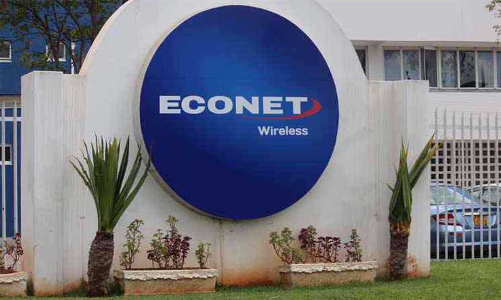 A round blue Econet Wireless sign on a white wall outside an Econet building, with palms and shrubs in front
